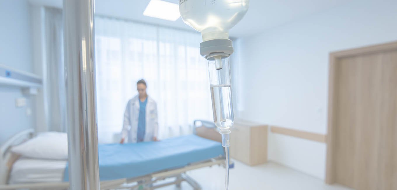 Капельница крупным планом, сзади медсестра застилает кровать